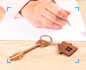 Primo piano di una mano che firma un documento su un tavolo in legno con una chiave di casa e un portachiavi a forma di casetta in evidenza.